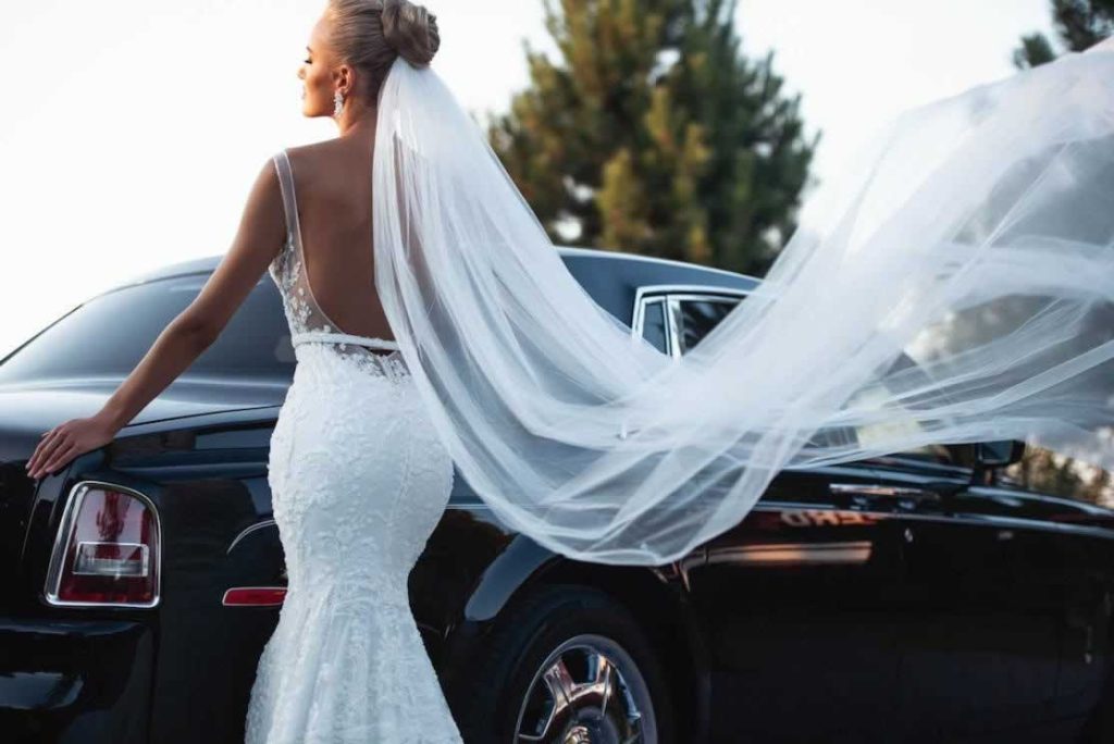 Rear view of a bride wearing a mermaid style lace wedding gown and veil in front of a black Rolls-Royce wedding car.
