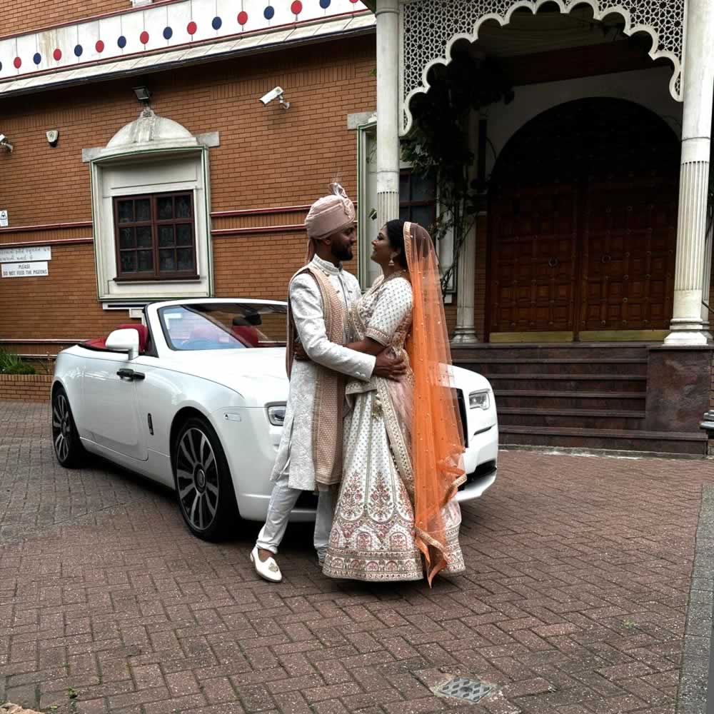Bride and groom wearing Indian wedding attire with Rolls-Royce Dawn convertible wedding car behind them