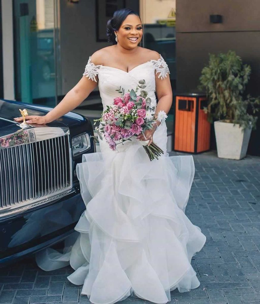 Bride wearing a lace trimmed off the shoulder sheath silhouette bridal dress with frilled skirt embellishments beside a black Rolls-Royce wedding car